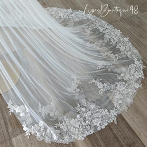May include: A white bridal veil with a floral lace trim. The delicate lace features detailed flower and leaf patterns. The veil is made of a sheer, flowing fabric, and the text "Lyns Boutique 98" is visible in the upper right corner.