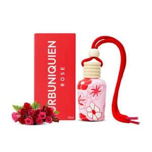 Op de afbeelding: Een roze luchtverfrisser met rode bloemmotieven en een rood touwtje. Een rode doos met de tekst "URBUNIQUIEN ROSE" en een klein boeket rode rozen staan ook op de afbeelding. De luchtverfrisser is 10 ml.