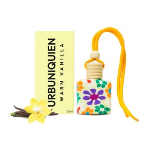 Op de afbeelding: Een luchtverfrisser met een bloemenpatroon, een gele koord en een houten kraal. De doos leest "URBUNIQUIEN WARM VANILLA 10ml". Een gele bloem en vanillestokjes zijn ook aanwezig.
