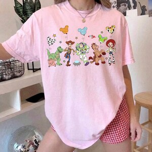 Puede incluir: Camiseta rosa claro con un gr&aacute;fico colorido de personajes de la pel&iacute;cula animada Toy Story. El dise&ntilde;o incluye a Woody, Buzz Lightyear, Jessie y otros personajes, junto con globos y estrellas. La camiseta es de cuello redondo y manga corta.