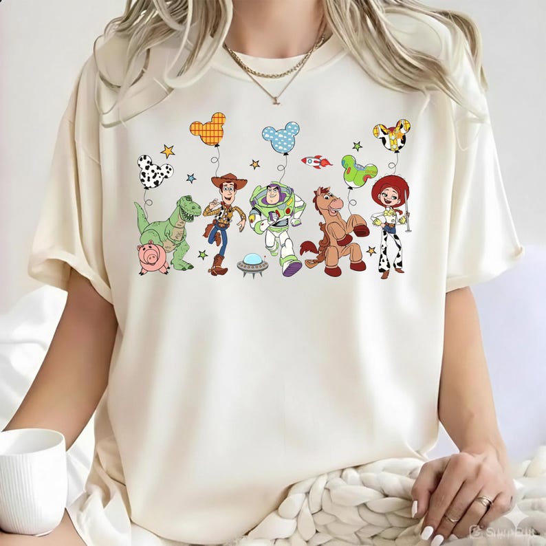 Puede incluir: Camiseta color crema con un gr&aacute;fico colorido de personajes de Toy Story, incluyendo a Woody, Buzz Lightyear y Jessie, con globos. El dise&ntilde;o incluye un dinosaurio verde, un cerdo rosa y un caballo marr&oacute;n. Los globos son de varias formas y colores.