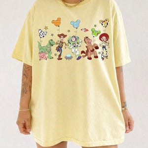 Puede incluir: Camiseta amarilla clara con un gr&aacute;fico colorido de personajes de dibujos animados y globos en forma de coraz&oacute;n. El dise&ntilde;o incluye un dinosaurio, un cerdo, Woody, Buzz Lightyear, un caballo y Jessie. La camiseta tiene mangas cortas y cuello redondo.