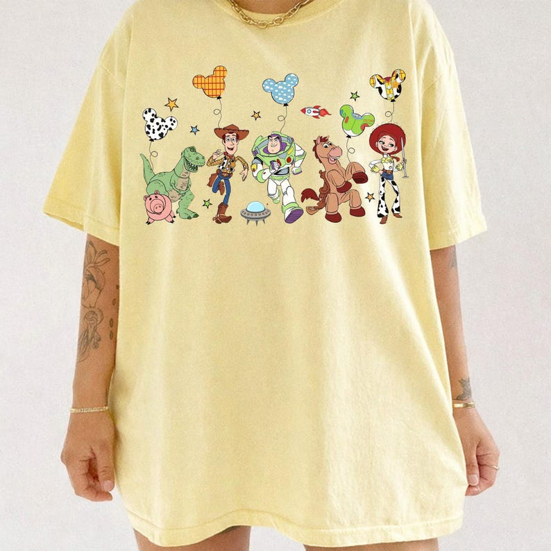 Puede incluir: Camiseta amarilla clara con un gr&aacute;fico colorido de personajes de dibujos animados y globos en forma de coraz&oacute;n. El dise&ntilde;o incluye un dinosaurio, un cerdo, Woody, Buzz Lightyear, un caballo y Jessie. La camiseta tiene mangas cortas y cuello redondo.
