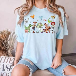 Puede incluir: Camiseta azul claro con un gr&aacute;fico colorido de personajes de Toy Story. El dise&ntilde;o incluye a Woody, Buzz Lightyear y otros personajes, junto con estrellas y globos. La camiseta es de estilo c&oacute;modo e informal.