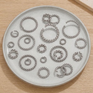 May include: An assortment of silver-toned, twisted metal earrings and ear cuffs presented on a pale grey ceramic plate. The earrings come in various sizes and shapes, including hoops and spiral designs. The jewellery is displayed on a neutral wooden surface.