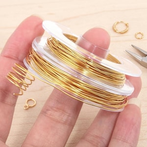 Peut inclure: Une bobine de fil doré, idéale pour la création de bijoux, est tenue dans une main. Plusieurs petits cerceaux dorés et un morceau de fil enroulé sont également visibles. Une pince est posée en arrière-plan.