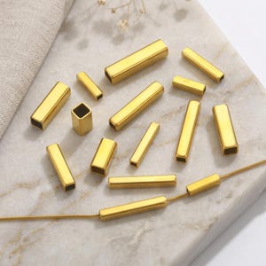 Puede incluir: Una colección de cuentas tubulares rectangulares de metal dorado con superficies lisas y centros huecos. Una cuenta está ensartada en una fina cadena de serpiente dorada, mientras que las otras doce cuentas están esparcidas alrededor en varias orientaciones.