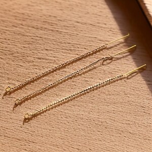 May include: Three delicate gold-colored earrings with chain and bar designs are displayed on a wooden surface. The earrings have a simple, elegant style, with a small loop at the bottom of each chain.