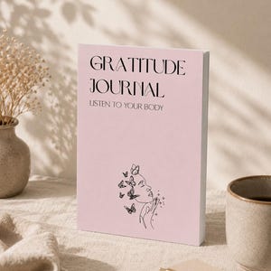 May include: A pink "Gratitude Journal" with the text "Listen to Your Body" and a line drawing of a face with butterflies. The journal is on a neutral-coloured surface with a pen, a mug, and a vase of dried flowers.