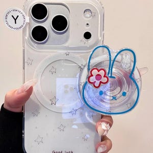 Op de afbeelding: Transparante telefoonhoes met een sterrenpatroon en een konijnvormige telefoonhouder. De houder heeft een blauwe omtrek en een rood-roze bloem. Op de hoes staat "Good luck".
