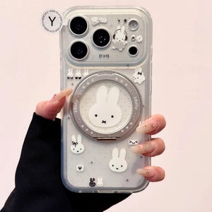Op de afbeelding: Transparante telefoonhoes met een Miffy konijn ontwerp en een zilveren ringhouder. De hoes heeft een lichtgrijze achtergrond met kleine konijn- en strik illustraties. De hoes heeft ook een camera bump met een konijn en ster ontwerp.