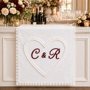Puede incluir: Un camino de mesa rectangular blanco con un corazón central delineado con ribete de tela blanca con volantes y un borde de encaje. Las letras C y R están bordadas en una fuente cursiva de color burdeos dentro del corazón.