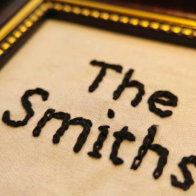 May include: Close-up of a framed embroidered piece with the words "The Smiths" in black thread. The embroidery is on a cream-colored fabric, set within a dark brown frame with a gold inner edge. The focus is on the handcrafted text.