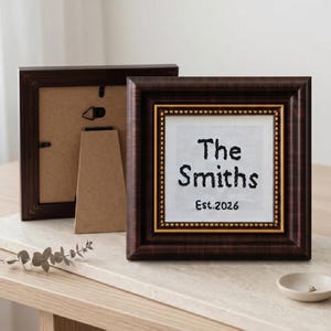 May include: Two brown wooden picture frames. The front frame has a white background with the text "The Smiths Est.2026" in black. The frame has a gold inner border and a dark brown outer border. The second frame is behind the first.