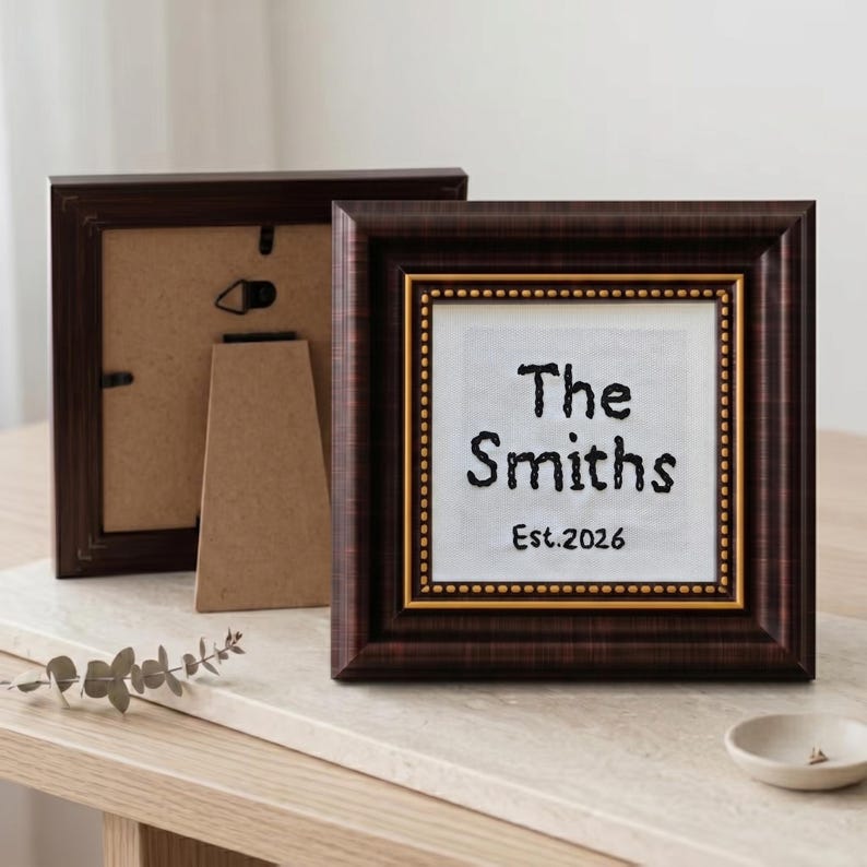 May include: Two brown wooden picture frames. The front frame has a white background with the text "The Smiths Est.2026" in black. The frame has a gold inner border and a dark brown outer border. The second frame is behind the first.