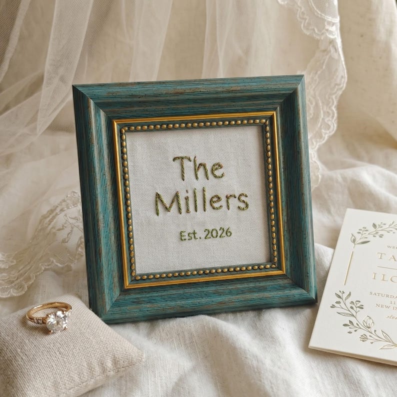 May include: A teal and gold framed embroidered piece with the words "The Millers Est. 2026". A gold ring with a large diamond sits on a small pillow. A wedding invitation is also visible.