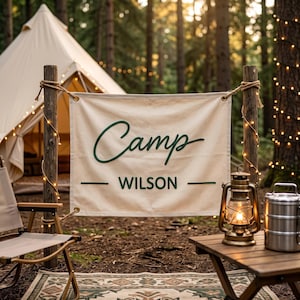 Könnte beinhalten: Ein rechteckiges, cremefarbenes Banner aus Segeltuch, das zwischen zwei Holzpfosten gespannt ist und den dunkelgrünen Schriftzug "Camp" in Schreibschrift über dem Wort "WILSON" in fetter serifenloser Schrift zeigt, flankiert von zwei Linien.