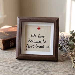 May include: A framed embroidery with the text "We love Because he first loved us." The artwork features a red cross and is set in a dark brown wooden frame. A vintage Holy Bible and a bundle of dried lavender are in the background.
