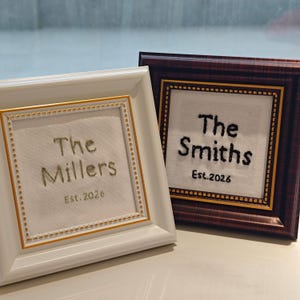 May include: Two framed embroidered pieces. One frame is white with gold trim, displaying "The Millers Est. 2026" in green thread. The other is a dark brown frame with gold trim, displaying "The Smiths Est. 2026" in black thread.