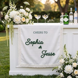 May include: A white fabric table runner with lace trim featuring the dark green text "CHEERS TO Sophia & Jesse" in a mix of serif and script fonts. The runner is displayed on a white bar with floral arrangements and glass bottles.