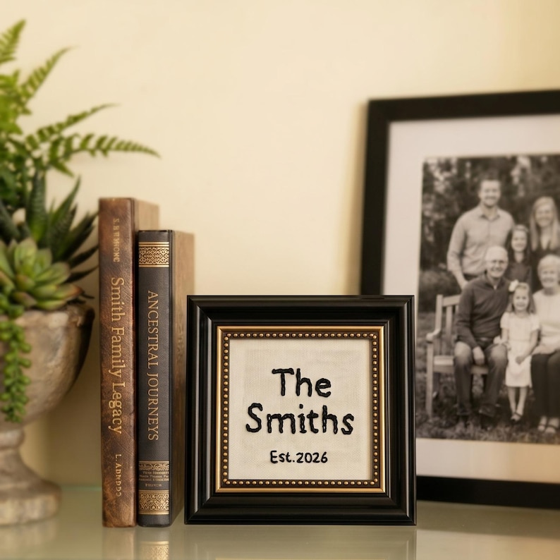 May include: A framed family name sign with the text "The Smiths Est. 2026" in black script. The frame is black with a gold inner border. Two antique books and a family photo are in the background.