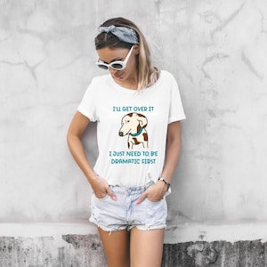 May include: White t-shirt with a cartoon dog graphic and the text "I'll get over it, I just need to be dramatic first." The dog has brown spots and a blue collar. The shirt is a casual, everyday wear item.