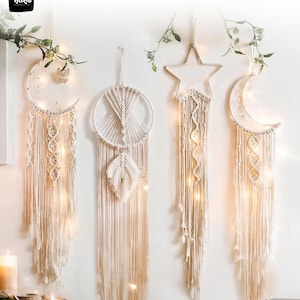 May include: Four macrame wall hangings in the shape of a moon, a circle, a star, and a crescent moon. The hangings are cream-colored with intricate knotting and fringe details. Fairy lights are woven throughout, and greenery is placed above.