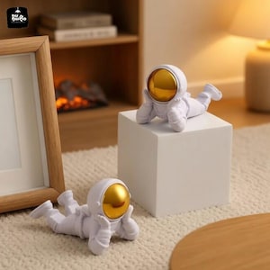 May include: Two white astronaut figurines with gold visors are displayed. One rests on a white cube, the other on a textured surface. The figurines are in a relaxed pose, suggesting a decorative or novelty item.