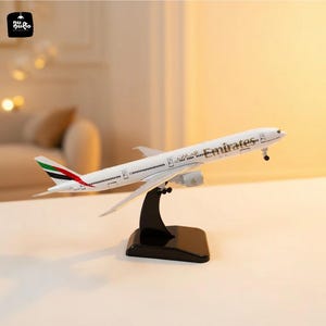 May include: A white Emirates airplane model with red, green, and black accents on the tail. The model is mounted on a black stand. The airplane has the Emirates logo and text printed on the side.