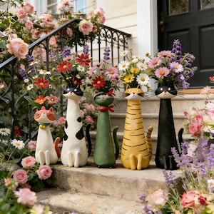 Peut inclure: Cinq vases en céramique en forme de chat, chacun contenant un bouquet de fleurs colorées. Les chats sont peints dans différentes couleurs et motifs, dont orange, noir et blanc, vert et jaune. Les vases sont disposés sur des marches extérieures.