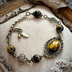 May include: An antique-style bracelet featuring tiger's eye stones and ornate bronze-toned metalwork. The bracelet has a central oval stone and several round stones, connected by delicate chain links and decorative filigree elements.