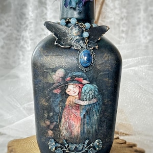 May include: A dark-toned decorative bottle featuring a central illustration of a person embracing a large blue bird. The bottle is adorned with a dark moth-shaped ornament, a blue oval gemstone pendant, and a sculpted leaf motif at the base.