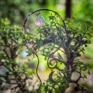 May include: A copper-colored wire plant trellis featuring a circular top adorned with clear faceted crystal beads, a small silver-toned star charm, and a hanging iridescent crystal sphere. A crescent moon charm hangs from a spiral at the base.