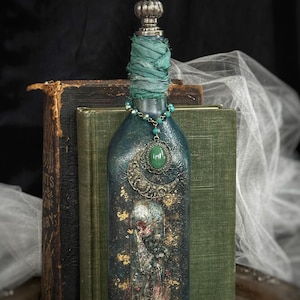 Puede incluir: Una botella de vidrio azul verdoso oscuro con un tapón de metal plateado y tela azul verdoso envuelta en el cuello. La botella está decorada con un dije de media luna, un colgante ovalado verde y una mariposa de metal con pan de oro.
