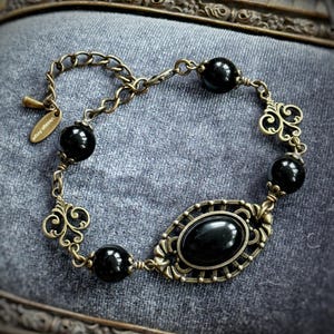 May include: An antique-style bracelet with a bronze chain and decorative elements. The bracelet features black, round beads and an oval, black centerpiece. The tag reads "Vintage Angel".