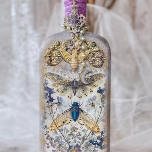 May include: A decorative glass bottle featuring a textured, light-colored finish adorned with gold-toned bee and insect embellishments, floral illustrations, a purple crystal stopper, and a dangling charm with a crescent moon and pearl-like beads.