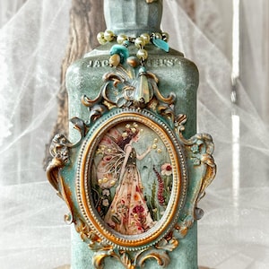 May include: A light green textured bottle featuring an oval frame with a fairy illustration. The bottle is adorned with a decorative metal topper, a beaded necklace with turquoise and pearl accents, and ornate gold-toned filigree embellishments.