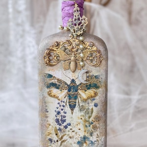 May include: A decorative glass bottle with a textured beige finish, featuring a central moth illustration, floral motifs, and gold filigree accents. The neck is wrapped in purple fabric, topped with a purple crystal, and adorned with a pearl and moon charm.