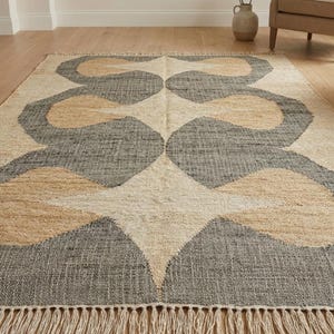 Puede incluir: Alfombra rectangular con un diseño geométrico en tonos neutros. La alfombra presenta un fondo crema con grandes formas abstractas en gris y beige. Tiene un borde con flecos y está colocada sobre un suelo de madera.