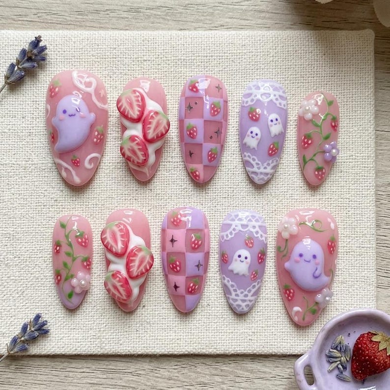 K&ouml;nnte beinhalten: Ein Set aus zehn mandelf&ouml;rmigen Press-on-N&auml;geln in Pastellrosa und Violett. Die Designs umfassen 3D-Erdbeerscheiben mit wei&szlig;er Creme, wei&szlig;e Spitzenmuster, kleine rote Erdbeeren sowie hellviolette Geister mit freundlichen Gesichtern.