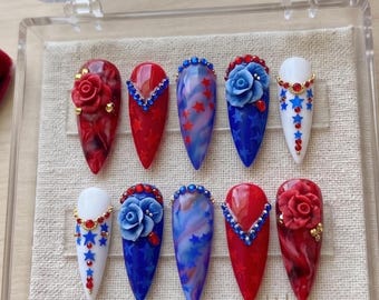 Pode incluir: Um conjunto de dez unhas artificiais com designs patrióticos. As unhas apresentam cores vermelhas, brancas e azuis, com enfeites como rosas, estrelas e strass. As unhas são exibidas em uma caixa de plástico transparente.