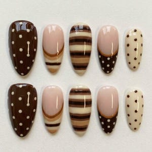 May include: A set of ten almond-shaped press-on nails featuring a brown and cream color palette. The designs include dark brown with white polka dots, nude with brown French tips, horizontal brown stripes, and cream with brown polka dots.