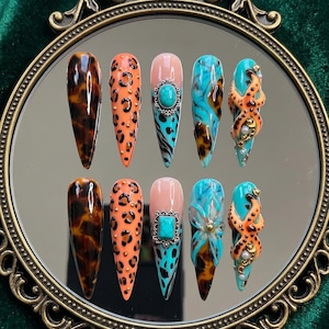 May include: A set of ten long, pointed artificial nails. Designs include tortoiseshell, leopard print, turquoise accents, and an octopus-like design with gold and pearl embellishments. The nails are arranged on a reflective surface.