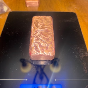 May include: A copper-colored rectangular bar with a textured surface, resting on a black digital scale. The scale displays a blue screen with numbers. Another similar bar is visible in the background.