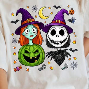 May include: A cream-colored t-shirt featuring a graphic of two characters wearing purple witch hats. One character has orange hair and a green pumpkin body, while the other is a skeleton with a black suit. Includes bats, webs, candy, and a moon.