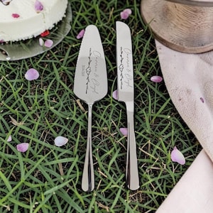 May include: A silver cake server and knife set engraved with the names "Carmen & Lloyd" and the date "26.06.2021". The set rests on green grass, surrounded by pink flower petals. A cake is partially visible in the background.