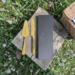 Könnte beinhalten: Goldfarbenes Tortenheber- und Messerset mit eingravierten Namen und Datum, präsentiert mit einer schwarzen Box. Das Set liegt auf einer rustikalen Holzoberfläche, umgeben von Grün und Blumen.