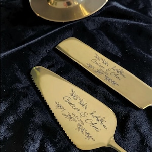 Może przedstawiać: Złoty zestaw do serwowania ciasta i szpatułki z wygrawerowanym tekstem i kwiatowymi akcentami. Tekst brzmi "Gulzer & Ami" i "05.04.2026". Zestaw jest prezentowany na ciemnoniebieskiej aksamitnej powierzchni, z złotym stojakiem w tle.