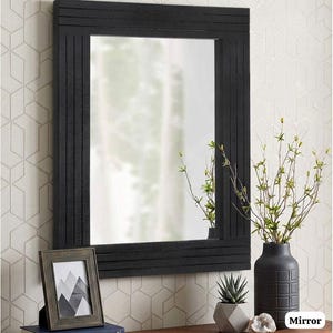 May include: A rectangular wall mirror featuring a black frame with vertical and horizontal grooved lines. The mirror reflects a bright interior space. The frame has a matte finish and a clean, structured design that surrounds the central glass.
