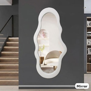 May include: A white, organically shaped wall mirror with a wavy, irregular frame. The mirror is mounted on a dark gray wall, reflecting a room with a chair and decorative items. The word "Mirror" is at the bottom.